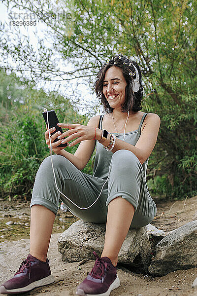 Lächelnde junge Frau benutzt Smartphone und hört im Park Musik über Kopfhörer
