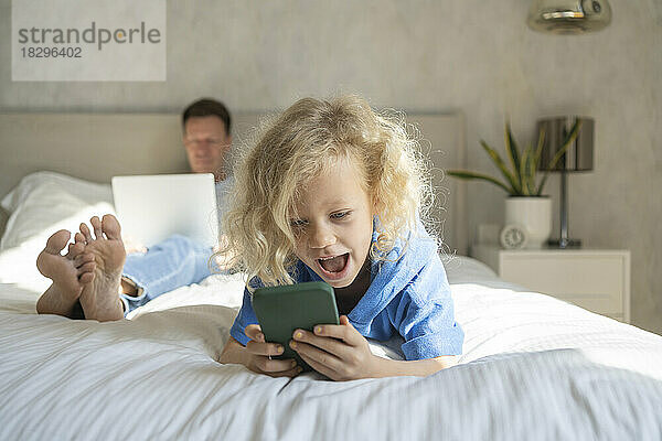 Glückliches Mädchen mit Smartphone und Vater mit Laptop im Hintergrund auf dem Bett