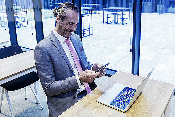 Glücklicher reifer Geschäftsmann  der Smartphone und Laptop am Schreibtisch im Büro nutzt
