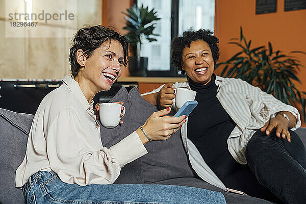 Glückliche Geschäftsfrauen mit Kaffeetassen sitzen auf dem Sofa im Büro