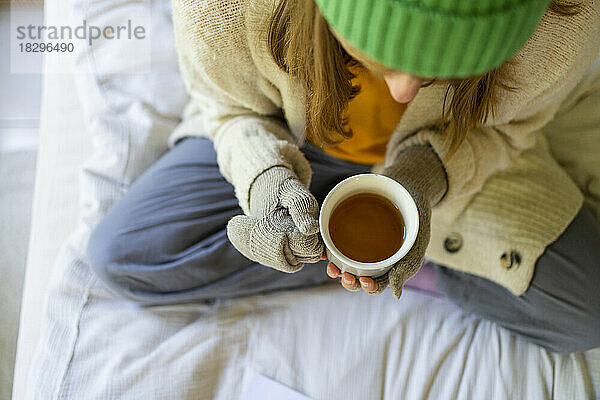 Frau mit Handschuhen sitzt mit Teetasse zu Hause im Bett