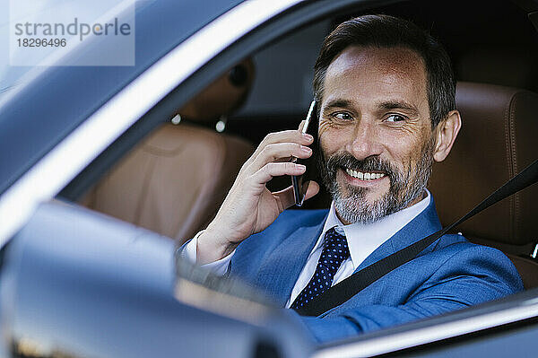 Lächelnder Geschäftsmann  der im Auto sitzt und über sein Smartphone spricht