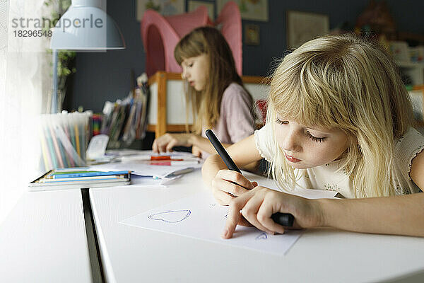 Mädchen mit Schwester zeichnet am Schreibtisch auf Papier