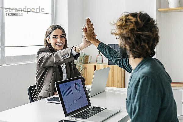 Lächelnde junge Geschäftsfrau gibt einem Kollegen am Schreibtisch High-Five