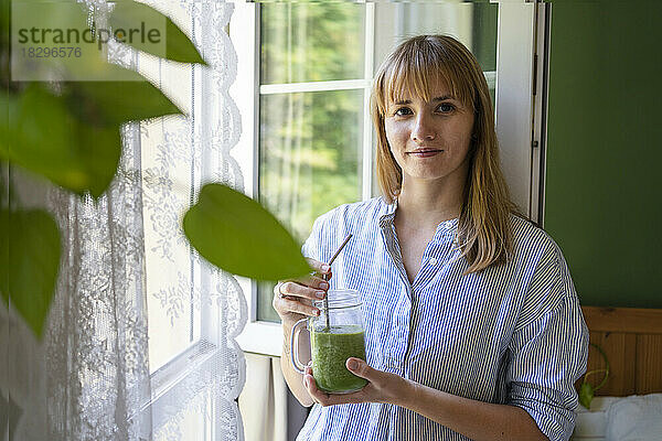 Lächelnde Frau hält Smoothie am Fenster