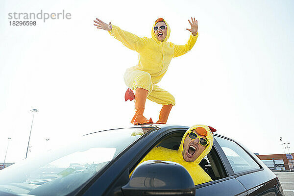 Mann schreit und fährt Auto  während unbekümmerte Frau im Hühnerkostüm auf dem Dach tanzt