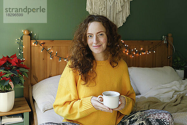 Lächelnde Frau hält zu Hause eine Tasse Tee auf dem Bett