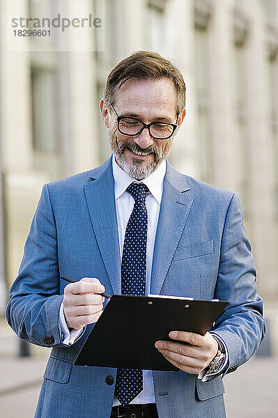 Smiling mature businessman standing with clipboard outside building
