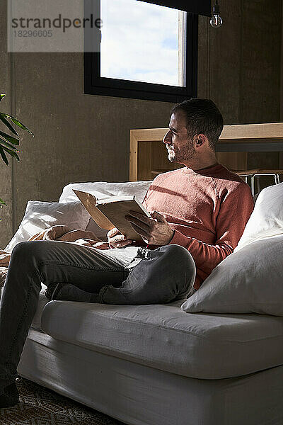 Nachdenklicher Mann sitzt mit Buch zu Hause auf dem Sofa