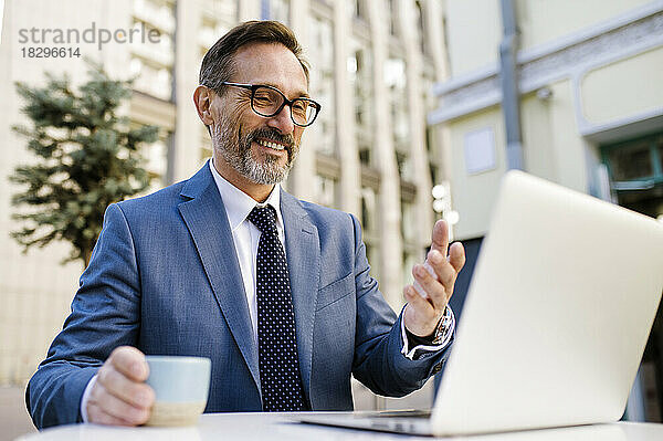 Lächelnder reifer Geschäftsmann mit Kaffeetasse gestikuliert bei Videoanruf über Laptop im Café