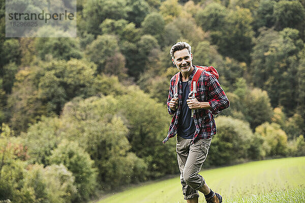 Lächelnder Mann mit Rucksack wandert auf Gras vor Bäumen