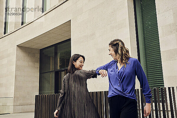 Glückliche junge Geschäftsfrau stößt vor dem Gebäude mit dem Ellenbogen gegen einen Kollegen
