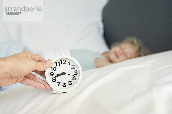 Hand of woman holding alarm clock in front of sleeping daughter on bed