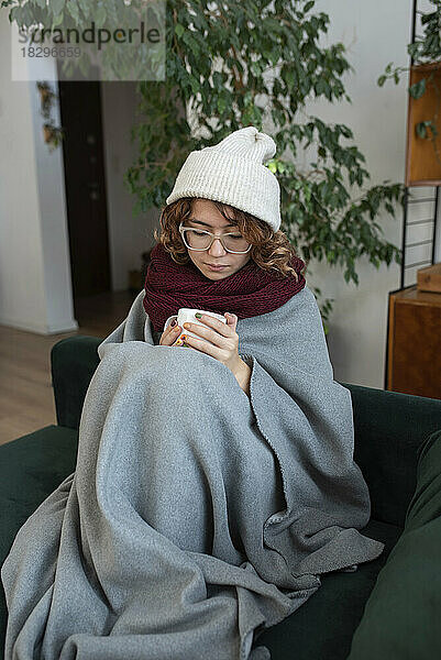 In eine Decke gehüllte Frau sitzt mit einer Tasse Tee zu Hause auf dem Sofa