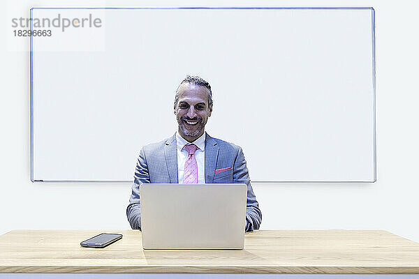 Lächelnder Geschäftsmann arbeitet am Laptop und sitzt vor dem Whiteboard im Büro