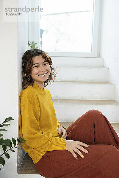 Lächelnde junge Frau sitzt zu Hause auf der Treppe
