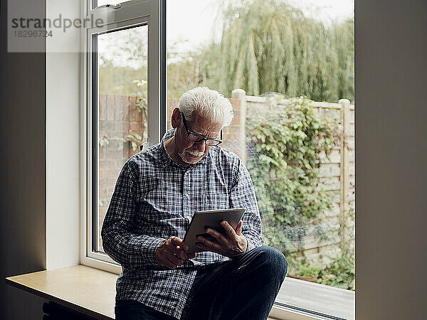 Älterer Mann am Fenster zu Hause mit digitalem Tablet