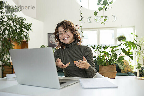 Glückliche junge Frau gestikuliert bei Videoanruf über Laptop zu Hause