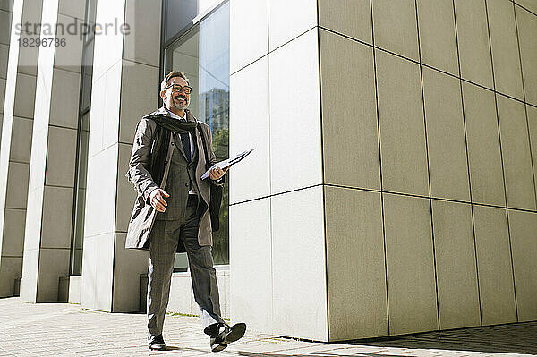 Lächelnder Geschäftsmann mit Klemmbrett  der auf dem Fußweg vor dem Bürogebäude läuft