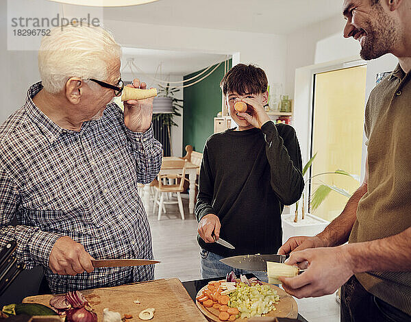 Großvater  Vater und Sohn schneiden gemeinsam in der Küche Gemüse und amüsieren sich mit Karotten