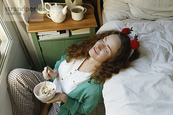 Lächelnde Frau mit geschlossenen Augen hält eine Müslischale neben dem Bett zu Hause