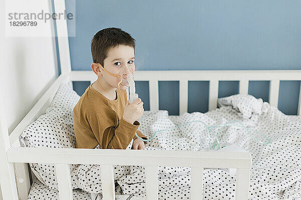 Junge mit Verneblermaske sitzt zu Hause im Bett