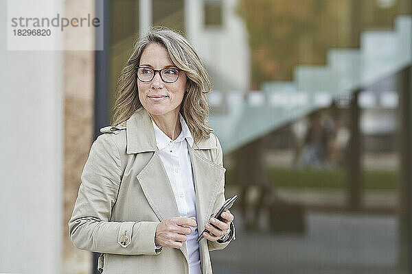 Nachdenkliche reife Geschäftsfrau steht mit Smartphone vor dem Gebäude