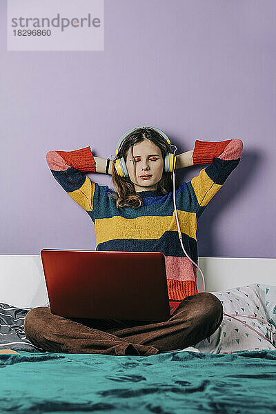 Mädchen mit den Händen hinter dem Kopf benutzt Laptop und Kopfhörer zu Hause im Bett