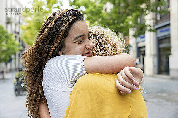 Smiling young woman with eyes closed embracing friend on footpath