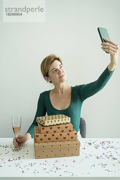 Lächelnde Frau macht ein Selfie per Smartphone mit einem Stapel Geschenke auf dem Tisch