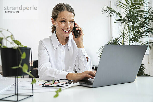 Lächelnde Geschäftsfrau  die im Büro mit dem Laptop telefoniert