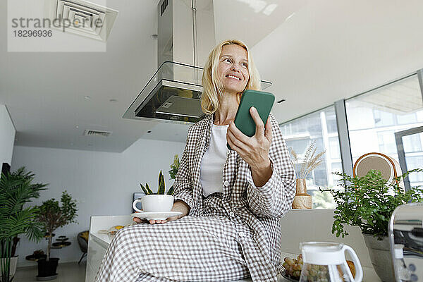 Lächelnde Frau mit Smartphone und Kaffeetasse sitzt auf der Küchentheke