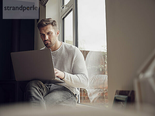 Mann am Fenster zu Hause mit Laptop