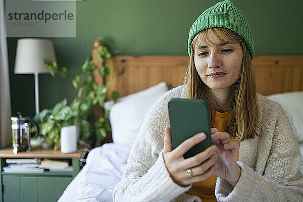 Lächelnde Frau mit Strickmütze und Smartphone zu Hause