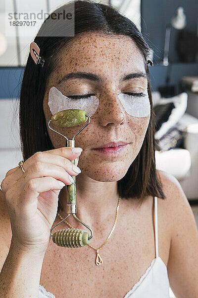 Frau mit Augenklappen massiert zu Hause mit einem Jadesteinroller ihr Gesicht