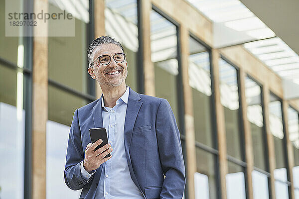 Fröhlicher reifer Geschäftsmann mit Mobiltelefon  der vor dem Gebäude steht