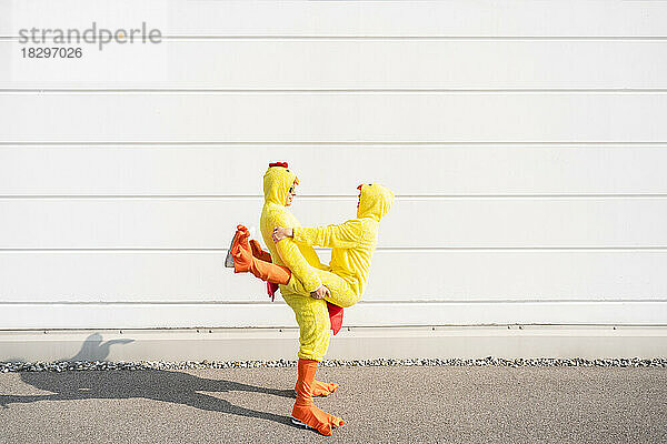 Verspielte Freunde in Hühnerkostümen stehen an einem sonnigen Tag an einer weißen Wand