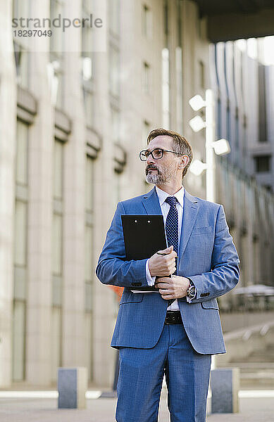 Thoughtful mature businessman with clipboard on footpath in front of building