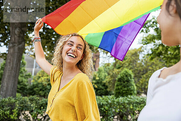 Glückliche Frau mit Regenbogenfahne und Blick auf Freundin im Park