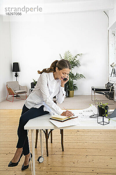 Lächelnde Geschäftsfrau  die am Schreibtisch im Büro sitzt und mit dem Mobiltelefon telefoniert und Notizen macht
