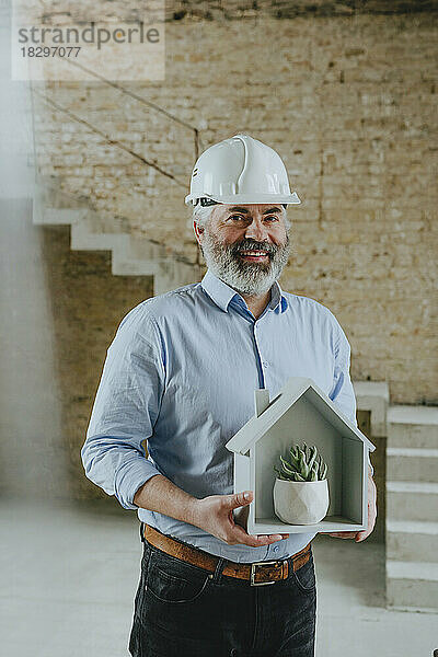 Glücklicher Geschäftsmann hält Hausmodell mit Topfpflanze auf der Baustelle