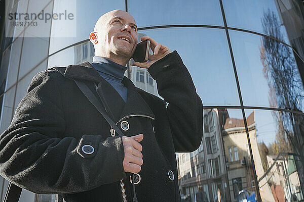 Lächelnder reifer Geschäftsmann  der vor dem Gebäude mit dem Smartphone spricht