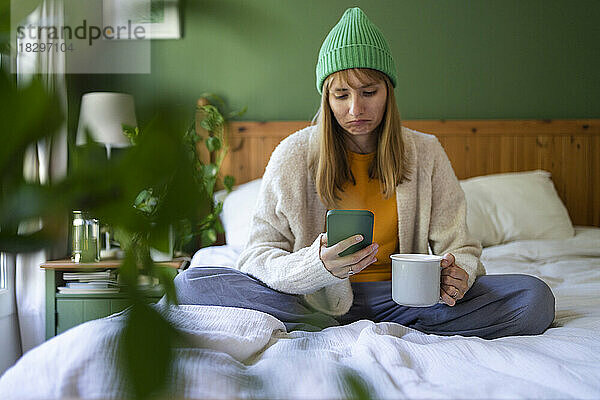 Traurige Frau benutzt Smartphone im Bett und trinkt zu Hause eine Tasse