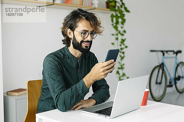 Glücklicher bärtiger Geschäftsmann  der sein Smartphone am Schreibtisch im Büro benutzt