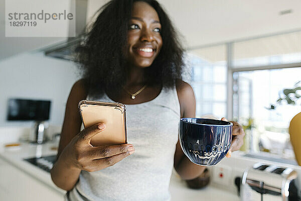 Lächelnde Frau hält Smartphone und Teetasse zu Hause