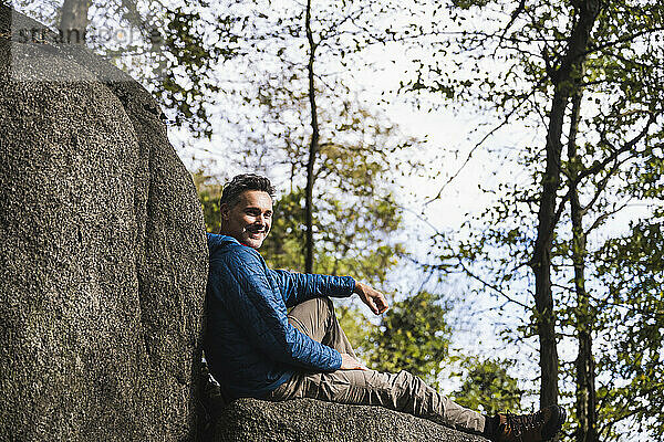 Lächelnder reifer Mann sitzt an einem Felsen vor Bäumen