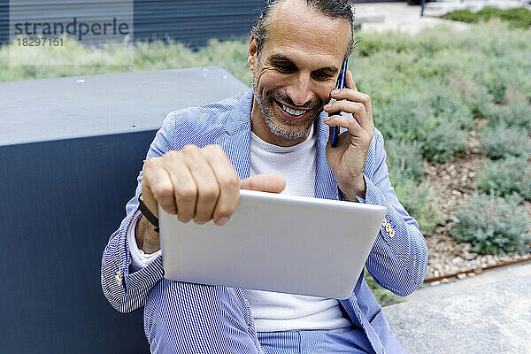 Glücklicher reifer Geschäftsmann  der auf einem Smartphone sitzt und einen Tablet-PC auf der Bank sitzt