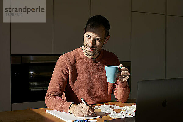 Lächelnder Mann mit Kaffeetasse  der am Schreibtisch auf Papier schreibt