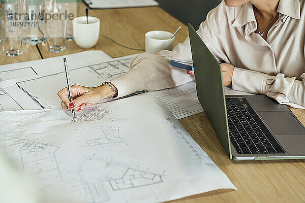 Businesswoman sitting with laptop and working on blueprint at table