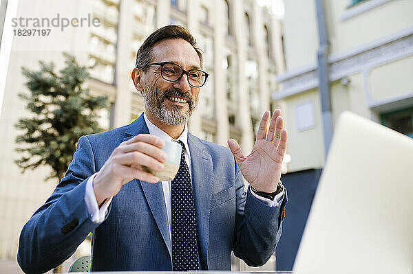 Glücklicher reifer Geschäftsmann  der im Café Kaffee trinkt und bei einem Videoanruf über seinen Laptop winkt